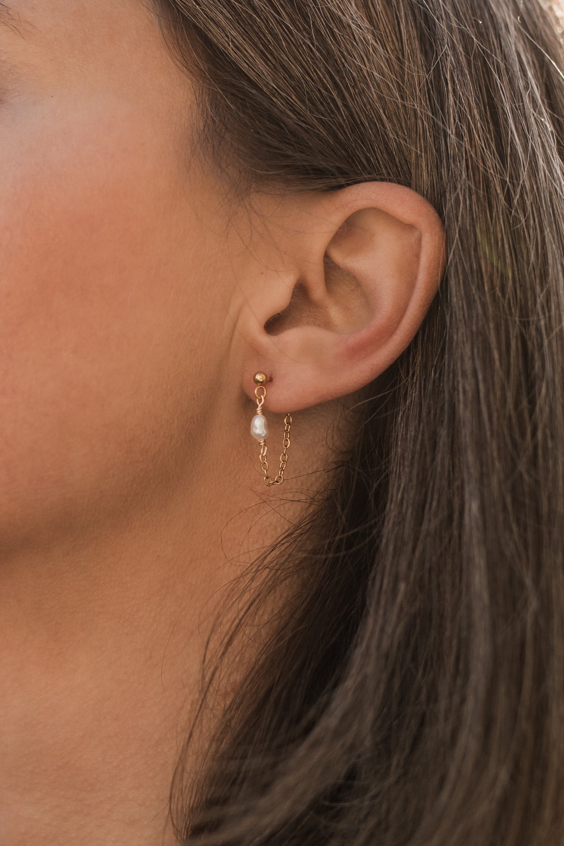 Detail řetízkových perlových náušnic v uchu modelky pro elegantní a minimalistický vzhled.