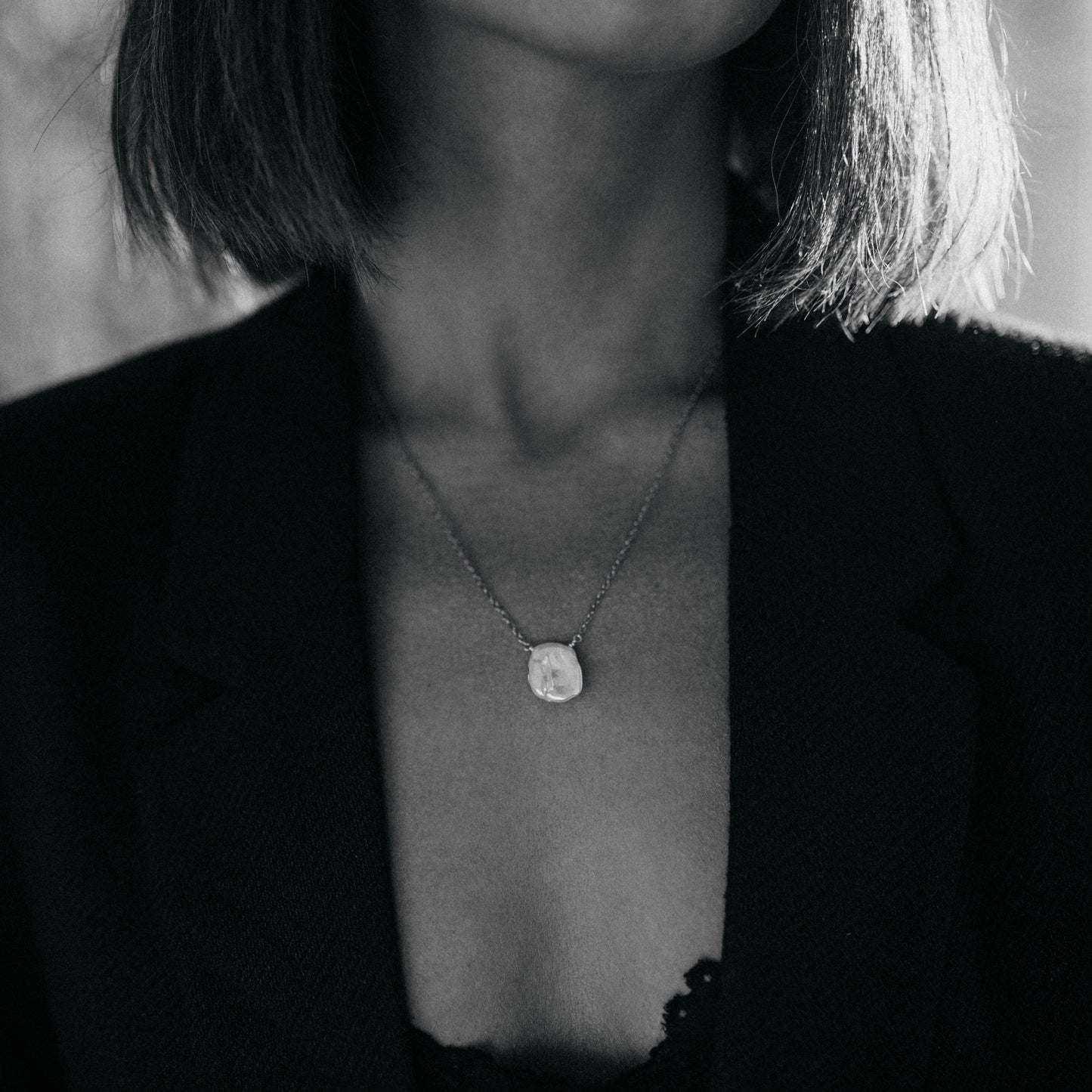 Detail jemného zlatého náhrdelníku s plochou keshi perlou na krku modelky, černobílá fotografie.