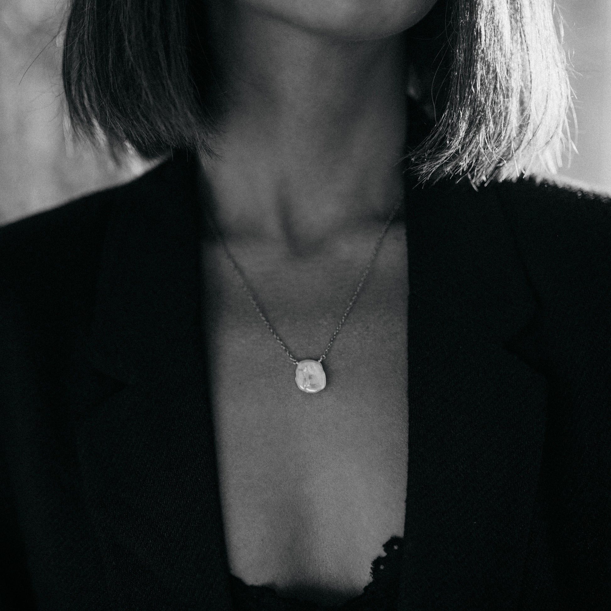 Detail jemného zlatého náhrdelníku s plochou keshi perlou na krku modelky, černobílá fotografie.